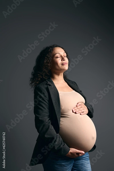 Fototapeta Delightful serene pregnant woman putting hands on her big belly, posing with her eyes closed over gray studio background