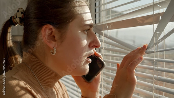 Obraz Portrait of stressed and scared woman loooking through window blinds on the street after seeing a crime