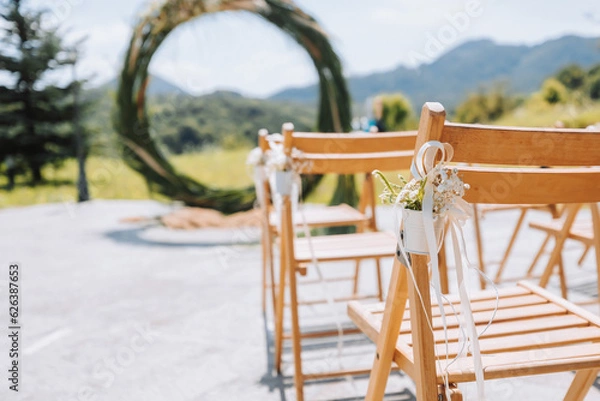 Fototapeta The wedding arch in the park is made of dry reeds on the background of the forest in blurred focus. Away wedding ceremony. Decorated chairs for the ceremony