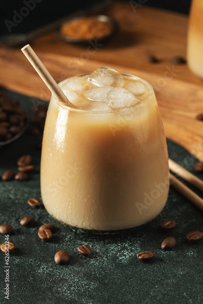 Fototapeta Ice coffee in a tall glass with cream poured over, ice cubes and beans on a dark concrete table.