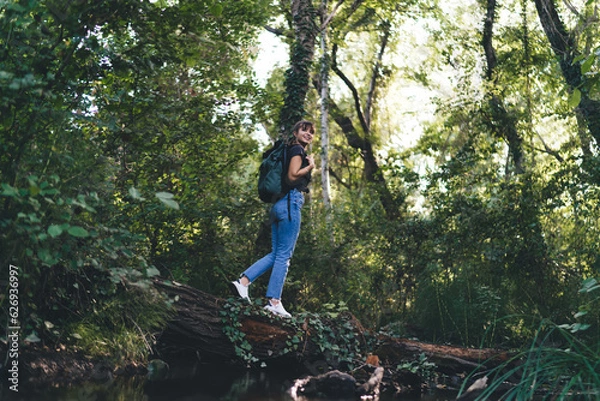 Fototapeta Trekking woman with backpack standing on tree trunk in forest