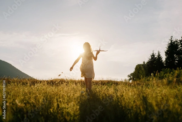 Obraz Silhouette of playful preteen girl kid run with airplane on background amazing summer warm sunset and majestic mountains. Dream freedom concept. Child runs on field holding in hands toy aircraft