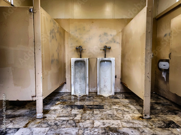 Fototapeta semi abandoned men's public toilet, very dirty and ruined, yellow light, dirty marble floor with various colors. Two urinals hanging on the wall, two other toilets on the sides