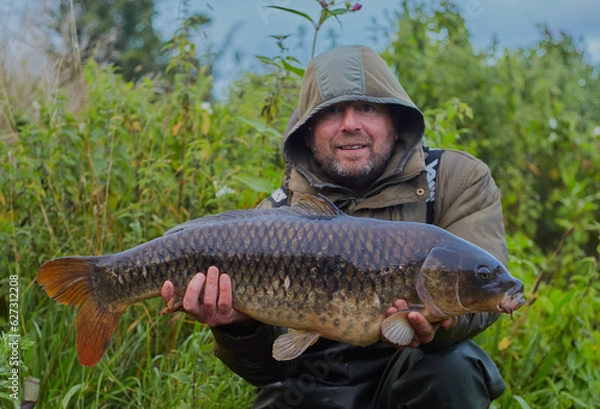 Obraz Fisherman holding up a common carp that he has caught with a smile on his face.