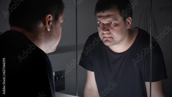 Fototapeta Upset and angry man in black t-shirt looking in his reflection at bathroom