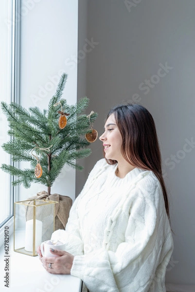 Fototapeta Woman in warm white winter sweater standing next to the window at home at christmas eve holding cup with marshmallows, fir tree behind