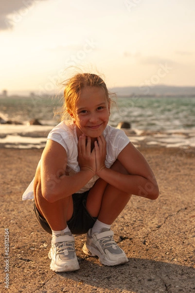 Fototapeta A girl dressed in a white dress is sitting on the pier. Smiling and looking ahead. The sea is restless and overcast before a thunderstorm