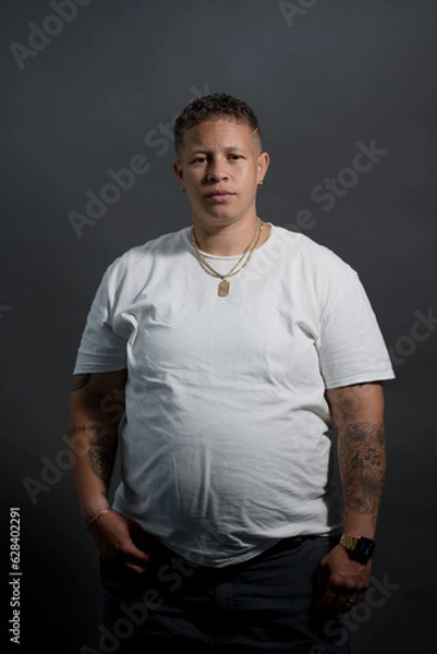 Fototapeta A woman in a white t-shirt poses for a casual portrait. She is in a studio, in front of a gray backdrop. Adding backstory, she is lesbian, with a butch aesthetic. She has a fade haircut and is Black.