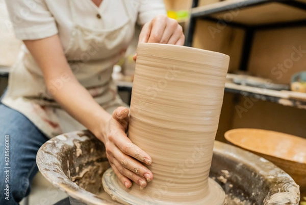 Obraz Cropped view of blurred craftswoman in apron creating clay vase on pottery wheel in workshop