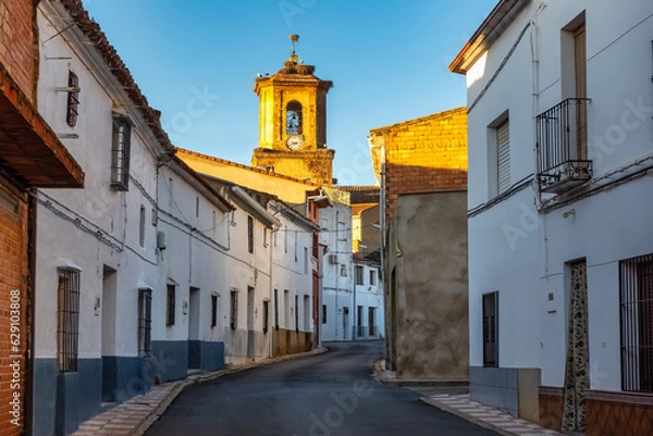 Obraz Alley with old houses painted white and tower of the Catholic church illuminated by the light of dawn, Agudo, Ciudad Real.