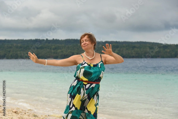 Obraz Happy attractive woman walks and dances on the beach. Senior woman wear the green long dress and sunglasses. Concept of active retired elderly and travel lifestyle. Landscape on the background.