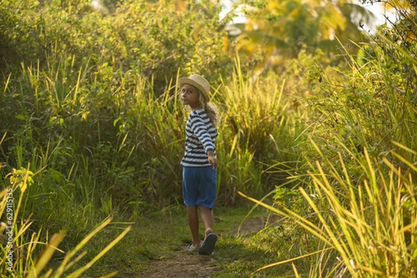 Obraz Rear view of blonde curly child 6 years old walking outside. The kid wears a hat, shorts and a blue-striped shirt. Happy childhood, travel. Palm trees in the background.