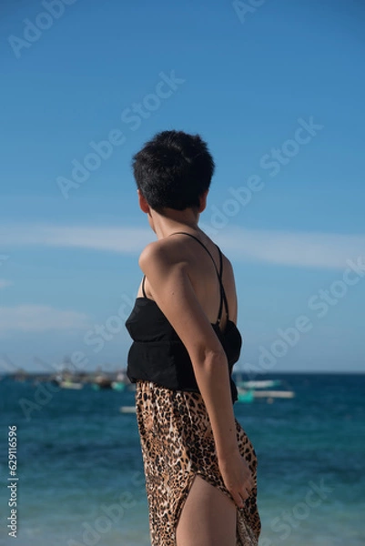 Fototapeta Young attractive girl walking at the sea beach in summer vacation. Rear view of stylish pretty woman with short black hair wears in leopard pant on the tropical beach with copy space.