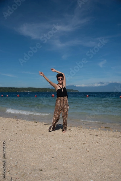 Obraz Young attractive girl standing near the ocean in summer vacation. Stylish pretty woman with short black hair wears in leopard pant having fun on the tropical beach. Copy space and blue sky.