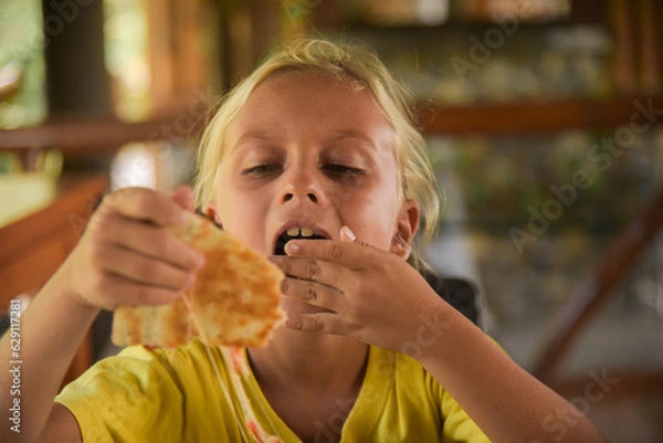 Obraz Adorable curly blonde boy eating pizza at the camera in italian restaurant. Portrait of child with long blonde hair and a yellow T-shirt eats junk food in a cafe. Blur on the background.