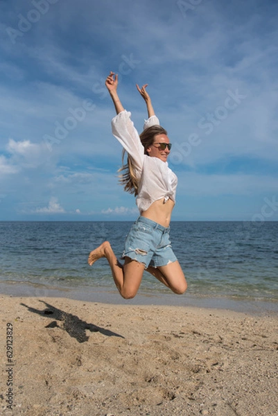 Obraz A happy blond woman with sunglasses is jumping on the beach. Attractive girl with long hair is wearing a denim shorts and a white shirt. Holidays and travel to other countries
