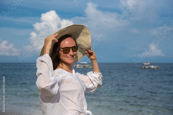 Obraz A happy blond woman with sunglasses is standing by the sea. She is touching her straw hat. A girl with long hair and a white shirt spends her vacation on the beach. Holidays to other countries