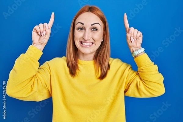 Fototapeta Young woman standing over blue background smiling amazed and surprised and pointing up with fingers and raised arms.