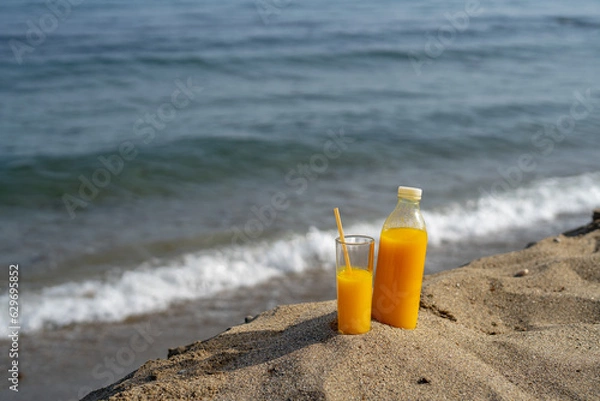 Fototapeta a bottle and a glass of orange juice stand on the sand against the background of the sea