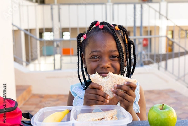 Fototapeta Afroamerykańska uczennica przy stole i zdrowy lunch z Sandwich w szkole podstawowej