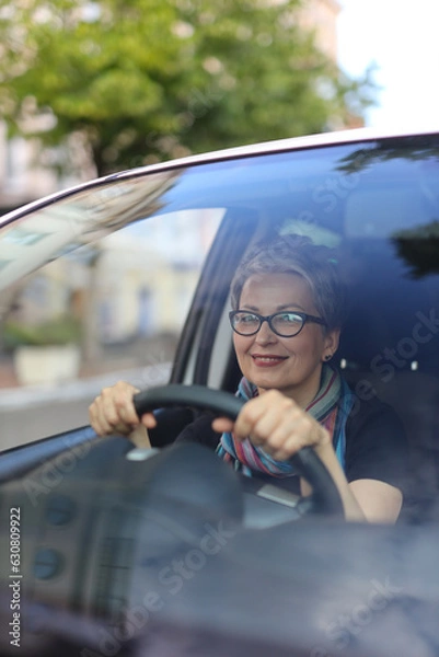 Fototapeta A senior businesswoman drives a new car on a summer day, enjoying the view and the trip. She is a successful and attractive owner of the vehicle.