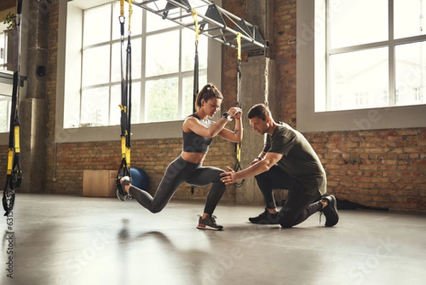 Obraz Doing squat exercise. Confident young personal trainer is showing slim athletic woman how to do squats with Trx fitness straps while training at gym.