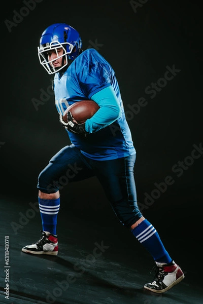 Fototapeta Portrait of a man in a blue uniform for american football runs with a ball on a black background. 