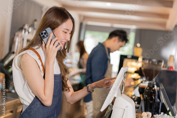 Obraz Asian coffee shop female employee accepts a pre-order on a mobile phone call while using a tablet computer in a cafe. Woman waiter talking on the cellphone. Home food delivery concept