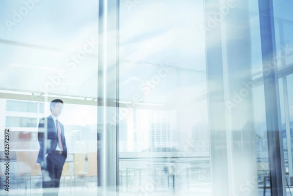 Fototapeta An Asian businessman standing in a suit looks out a glass window of an office building.  There are reflections on the glass.