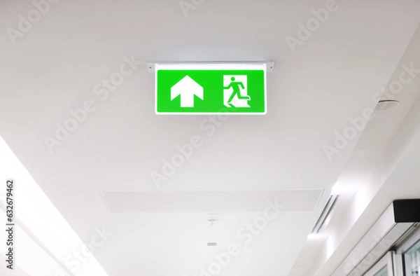 Fototapeta Lighted glowing green emergency exit signs in an office hallway with arrows pointing the way out of the building.