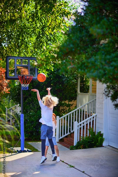 Fototapeta happy kids playing basketball at the driveway of their home. portable basketball hoop stand. active lifestyle. neighborhood activity sports