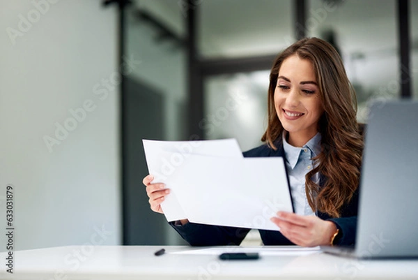 Obraz A smiling female employee is doing paperwork and using a laptop at the office.