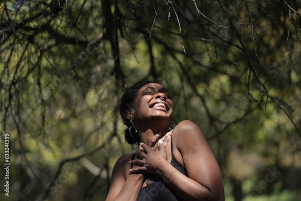 Obraz With their hands clasped at their chest and laughing joyfully, a self-described nonbinary person poses for a happy portrait. Adding narrative, the model is Black, and surrounded by nature.