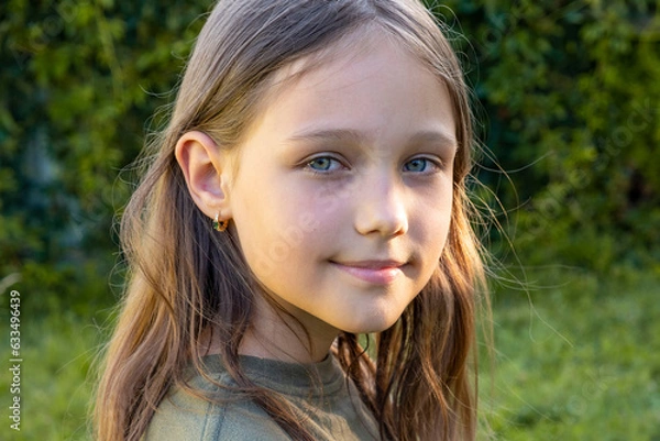 Obraz Portrait of a beautiful brunette little teenager girl looking at the camera and smiling, standing on the street against the backdrop of greenery on a summer day. Young curious child raises his eyes