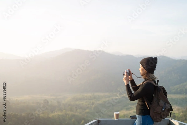 Fototapeta Hiker photographer taking picture of valley with mountains from view point. woman wearing hat stand alone and enjoying freedom and calm inspired travelling. Tourist traveler on background view mockup