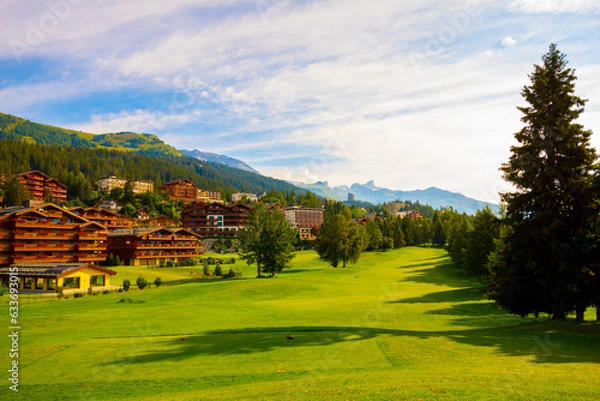 Fototapeta Village with House and Hotel with Panoramic View over Mountain with Clouds and Golf Course in a Sunny Summer Day in Crans Montana, Valais in Switzerland.