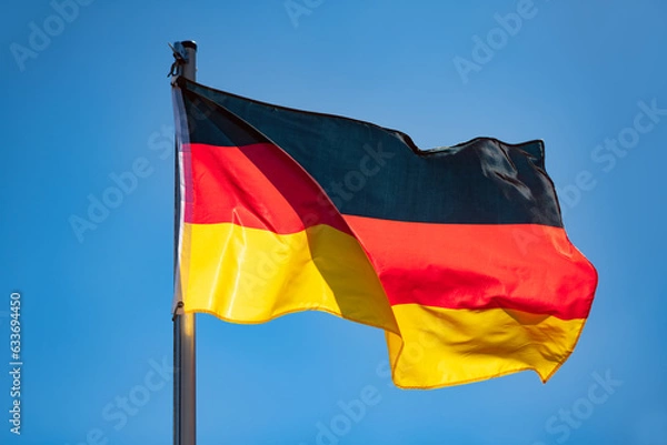 Fototapeta German flag in black-red-gold color on a sunny day in Germany. National yymbol of the “Bundesrepublik Deutschland“. Flag pole and wavy fabric blown in the wind on a blue sky day. Intense sunlit colors