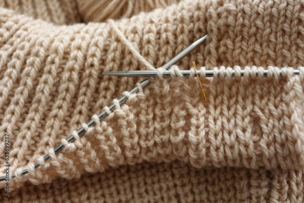 Fototapeta Knitting project in progress. Beige sweather not finished yet. Uneven loops and columns. Warm and comfort background with closeup knitting needles in work.