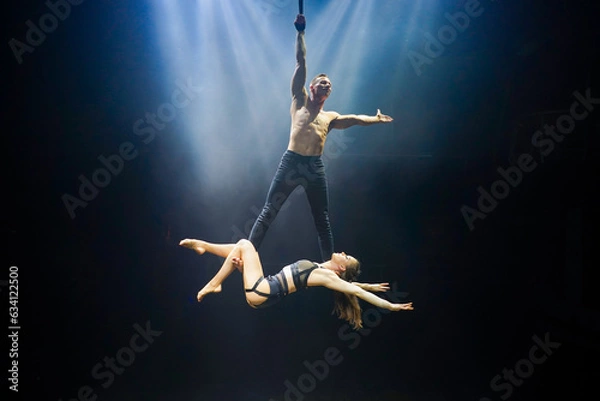 Fototapeta An acrobatic performance featuring an aerial straps duet: a man gracefully hangs by one hand, while a woman lies extended at his feet. They are bathed in an ethereal blue and white glow
