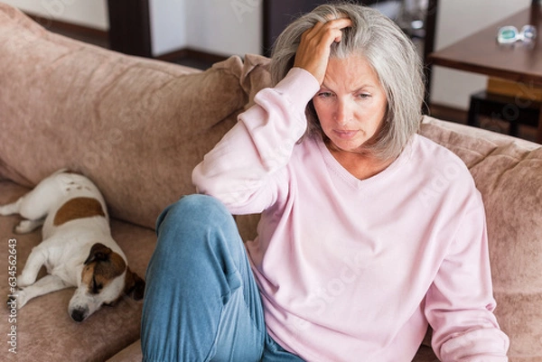 Fototapeta Sad pensive middle age woman looking down depressed sitting on the sofa