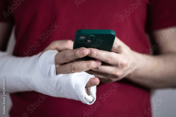 Fototapeta A man in a red T-shirt with a broken arm in a cast holds and uses a phone Close-up