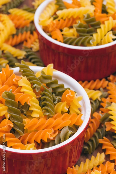 Obraz Three-colored uncooked pasta in the red ceramic bowls on the wooden background with scattered pasta on it.
