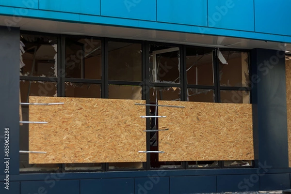Fototapeta Civil building damaged by military drones attack. Windows with broken glass covered with OSB boards. Repairing after war afthermath