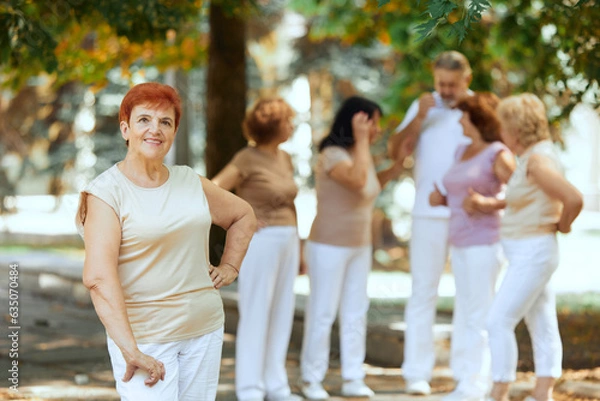 Obraz Focus on senior redhead smiling woman standing in park on warm sunny day. Blurred people on background. Concept of sport and health, active lifestyle, age, wellness, body, care