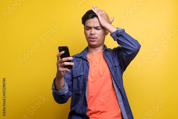 Fototapeta Shocked Asian man in casual clothes touch his head while looking at smartphone with dissapoint expression isolated on yellow background