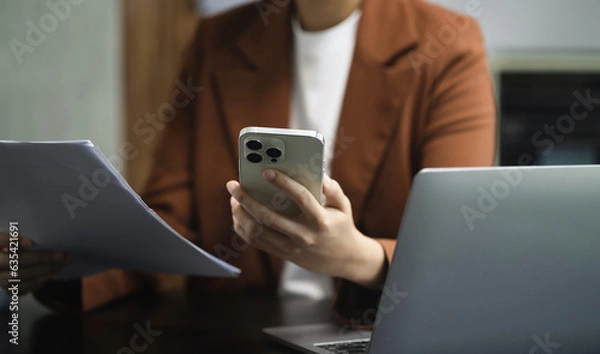 Fototapeta Employee, working with documents sitting at desk using laptop and smartphone at work.