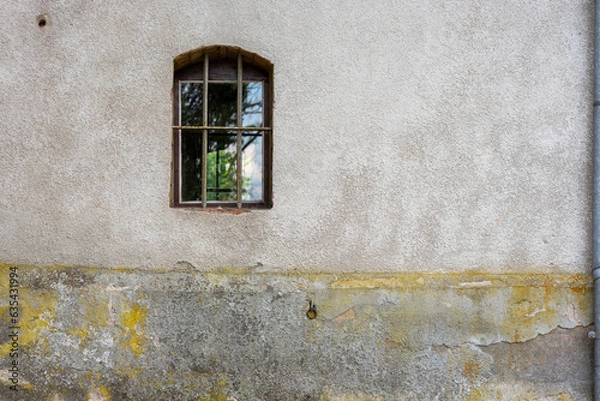 Fototapeta A small arched window protected by a lattice on a solid concrete wall that is weathered and faded.
