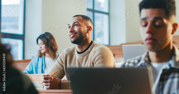 Fototapeta Handsome young African American male student sitting among others, listening to professor and noting information at lection. High school concept. Guy studying at University and writing down.