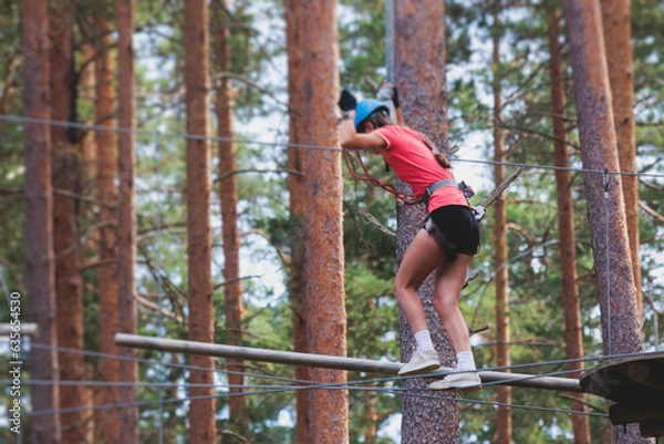 Fototapeta View of high ropes course, process of climbing in amusement acitivity rope park, passing obstacles and zip line on heights in climbing safety equipment gear between the trees om heights, summer day