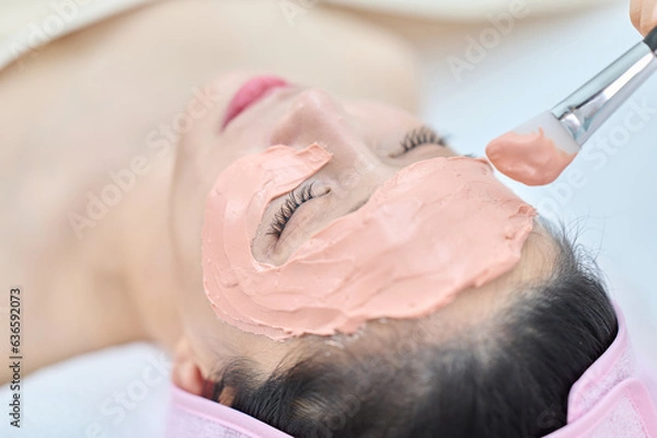 Fototapeta 아시아 한국의 젊은 여성이 스파 또는 피부관리전문점 또는 피부과 병원에서 분홍색 두건을 쓰고 침대에 누워 베이지색 대형 타올을 덮고 하얀색 셔츠를 입은 전문관리사가 실리콘붓으로 모델링팩을 얼굴에 바르고 있다. 